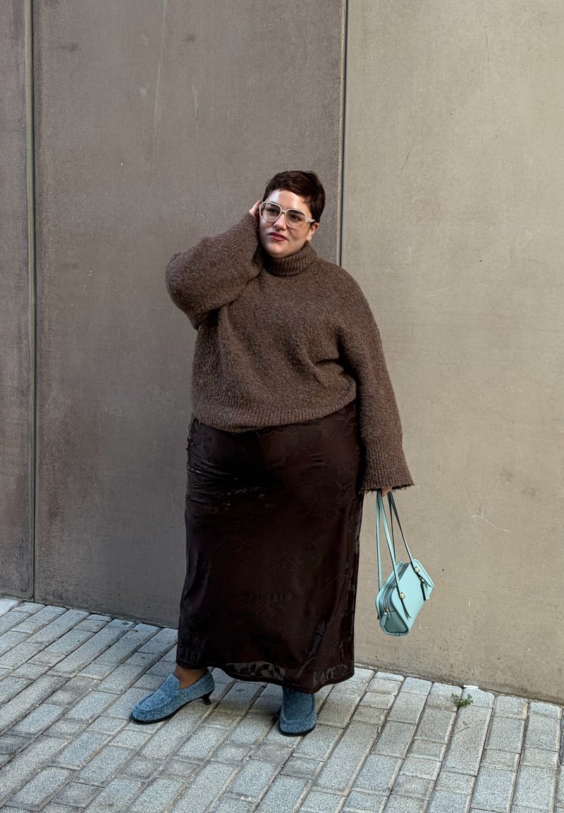 Personne aux cheveux courts portant un pull marron surdimensionné, une longue jupe foncée, des chaussures bleues, tenant un sac à main bleu clair, debout sur un trottoir carrelé près d'un mur en béton.