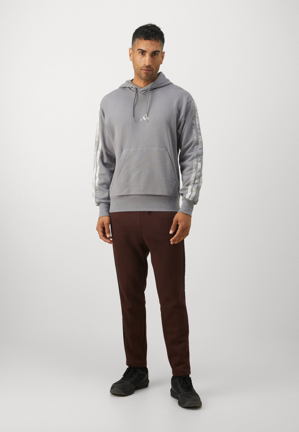 Sudadera con capucha gris, con un bolsillo frontal y mangas a rayas plateadas, combinada con pantalones de chándal marrones oscuros y zapatos negros.