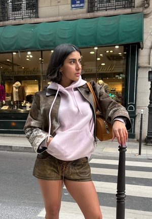 Chaqueta marrón con patrón, sudadera con capucha rosa claro, pantalones cortos verdes con cordones y bolso marrón. Ambiente urbano con un escaparate de fondo.