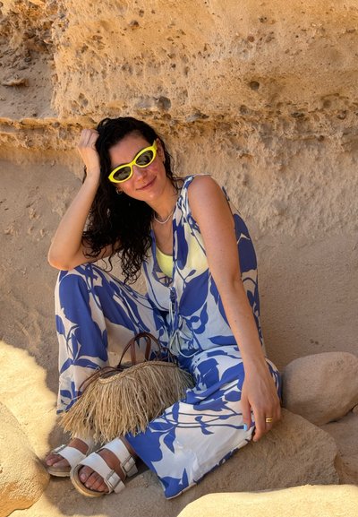 Mono de pierna ancha en azul con patrones florales blancos. Los accesorios incluyen un bolso de paja y sandalias blancas. Unas gafas de sol amarillas brillantes completan el look.