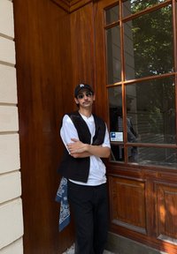 Gilet noir porté sur une chemise blanche, pantalon noir, et un bandana bleu à motifs suspendu. Fenêtre en verre et porte en bois foncé en arrière-plan.