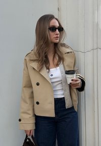 Beige színű, dupla gombos zakó nagy fekete gombokkal, fehér tank topra rétegezve. Sötét farmernadrág és fekete táska egészíti ki a megjelenést. Kávéscsészét tart a kezében.