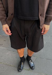 Juodi klostuoti šortai derinami su juoda marškinėliais ir rudais megztiniais. Blizgūs juodi loafersai papildo aprangą. Kasdienis stilius ant šaligatvio.