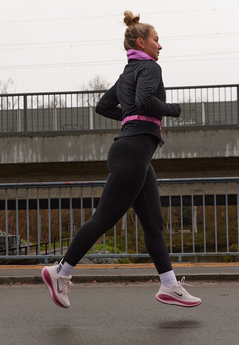 Coureuse en leggings noirs et veste noire avec des accents roses, portant des chaussures Nike roses avec des semelles blanches, courant en plein air sur un chemin.