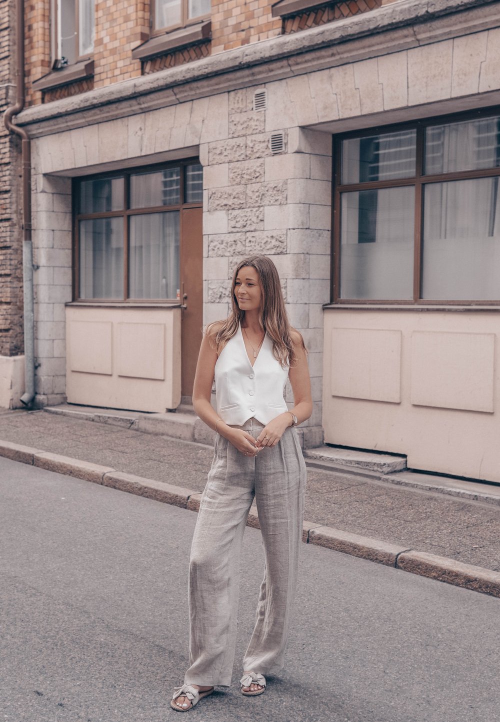 White sleeveless vest with front buttons, paired with light gray linen wide-leg pants and sandals, set against a brick building backdrop.