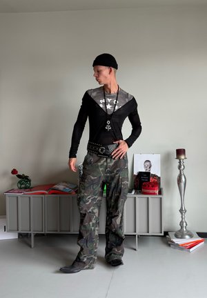 Camiseta negra de mangas largas con estampado de camuflaje gris, combinada con pantalones cargo de camuflaje. Accesorios incluidos un cinturón con tachuelas y un collar largo.