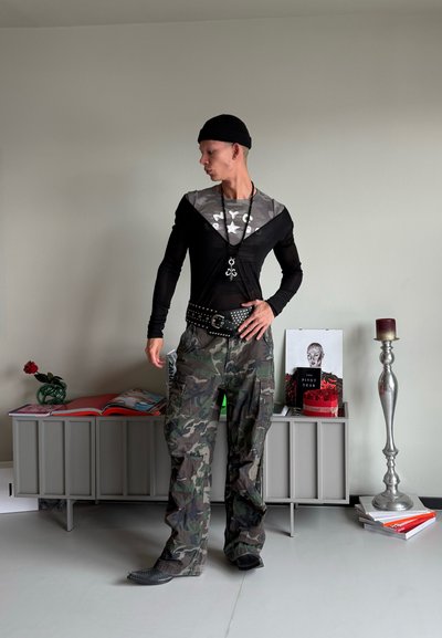 Camiseta negra de mangas largas con estampado de camuflaje gris, combinada con pantalones cargo de camuflaje. Accesorios incluidos un cinturón con tachuelas y un collar largo.