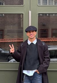 Egy személy fekete lapos sapkában, sötét pulóverben és fekete kabátban zöld kétszárnyú ajtók előtt áll, és békejelet mutat.