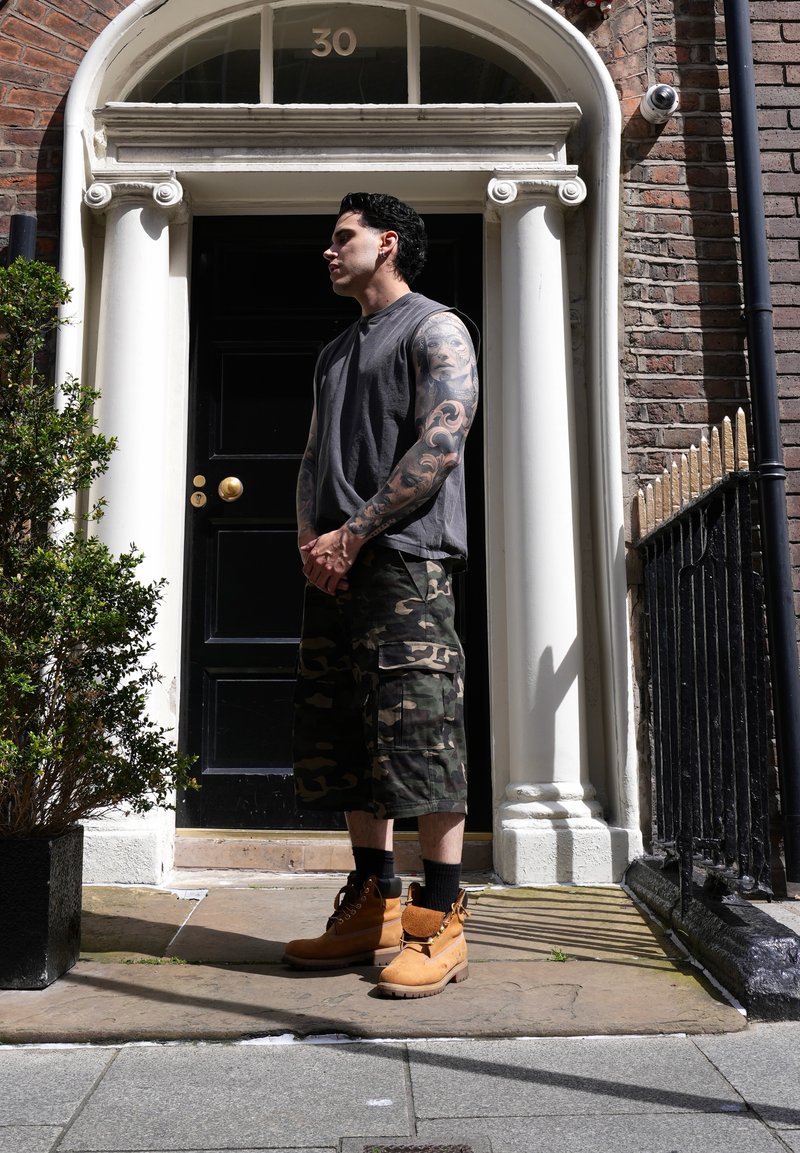 Homme au bras tatoué portant un débardeur, un short camouflage et des bottes, debout devant une porte noire numérotée 30 encadrée par des piliers blancs.
