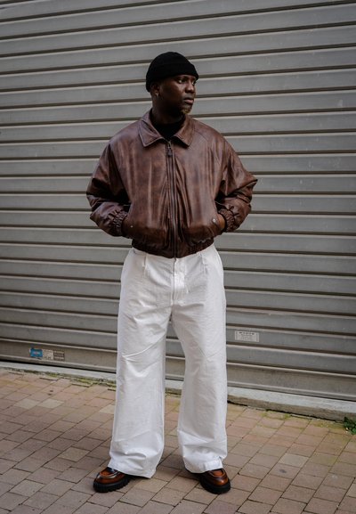 Chaqueta bomber de cuero marrón, pantalones anchos blancos, gorro negro, zapatos marrones y negros, de pie contra un fondo gris.
