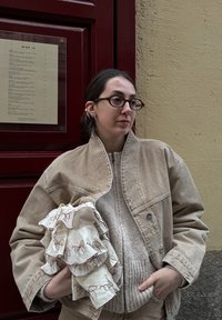 Bezs színű farmerdzseki egy texturált krém pulóver fölött. Egy fodros anyagot tart, amely vörös hímzéssel díszített. A háttérben egy bordó ajtó és fal látható.