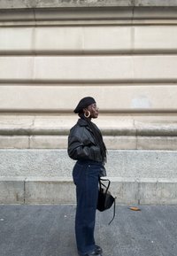 Zwarte leren cropped jas, donkerblauwe high-waisted denim jeans, zwarte baret, gouden creooloorbellen en ronde bril tegen een stenen muur.