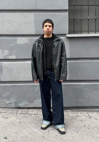 Νέος άνδρας με κοντά μαλλιά στέκεται μπροστά σε γκρίζο τοίχο φορώντας μαύρο δερμάτινο μπουφάν, μαύρο πουλόβερ, φαρδιά σκούρα τζιν γυρισμένα στις άκρες και μπεζ παπούτσια.