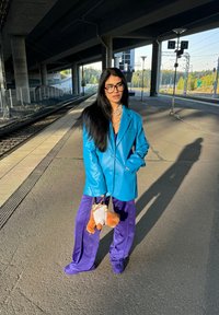 Blazer de cuero azul, pantalones morados de pierna ancha, gafas negras oversized, collares en capas y un pequeño bolso con un acento de oso de peluche.