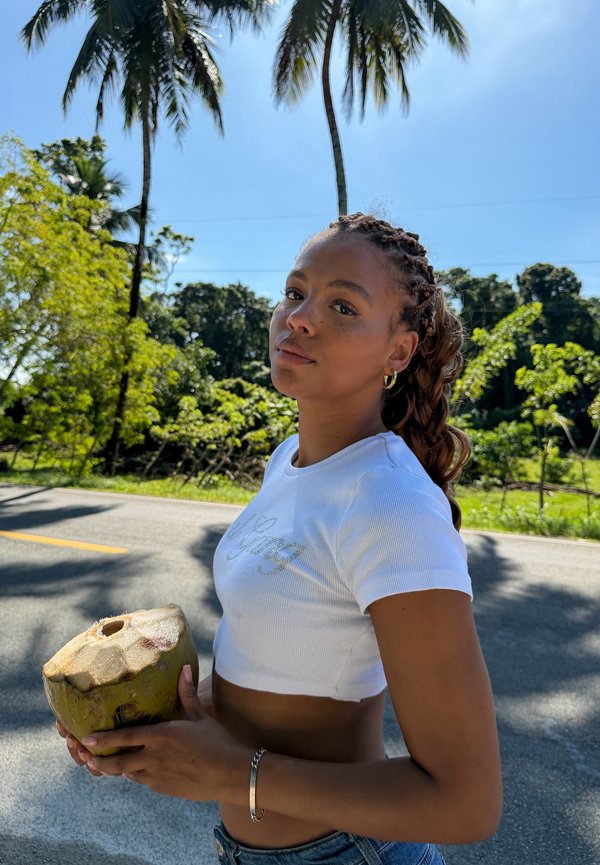 Jonge vrouw met gevlochten haar die een verse kokosnoot vasthoudt, staand langs een zonovergoten weg met palmbomen en groen op de achtergrond.