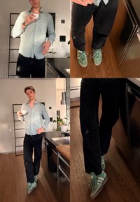 Νεαρός άνδρας με ριγέ πουκάμισο και μαύρο παντελόνι πίνει από ένα λευκό φλιτζάνι σε μια μοντέρνα κουζίνα, φορώντας πράσινα αθλητικά παπούτσια πάνω σε ξύλινο πάτωμα.