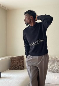 Man in zwarte trui met de tekst "Anthem Cotton" en grijze broek staat met één hand achter zijn hoofd in een kamer met een witte bank en patroonige kussens.