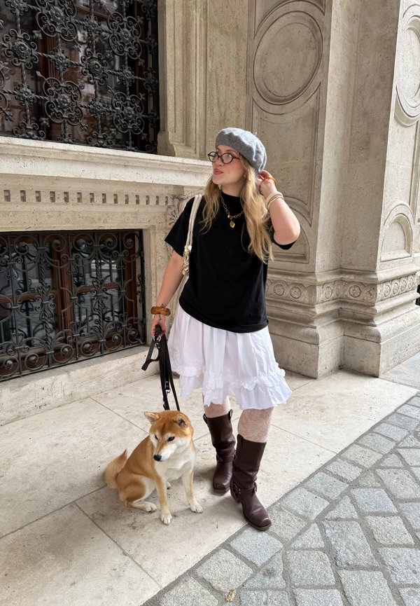 Uma pessoa veste uma t-shirt preta, uma saia branca em camadas e botas castanhas até a coxa. Ela segura um cão com uma coleira perto de uma parede decorativa.