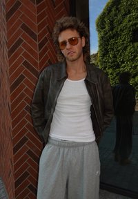 Brun læderjakke over en hvid top, parret med grå sweatpants. Accessories inkluderer brune solbriller. Baggrunden har murstenmønster.