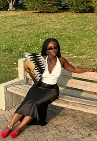 Jupe maxi en satin noir, haut halter blanc, éventail multicolore avec un motif géométrique, lunettes de soleil oversize, ballerines rouges, assise sur un banc en bois.