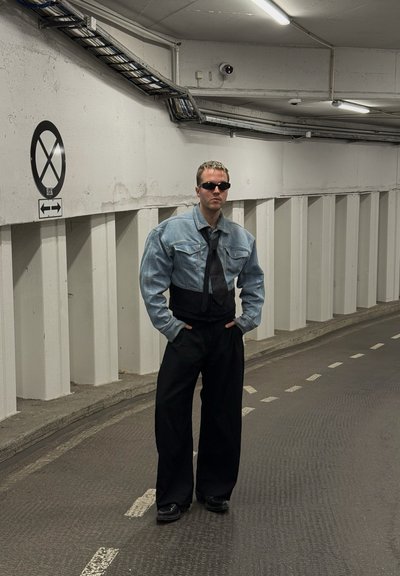 Chaqueta de mezclilla con hombros anchos, corbata negra y pantalones negros holgados. Llevada con gafas de sol, en un aparcamiento de hormigón.