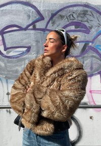 Faux fur jakke i lys brun, med en tæt, blød tekstur og oversize krave. Båret over en sort top med blå jeans.