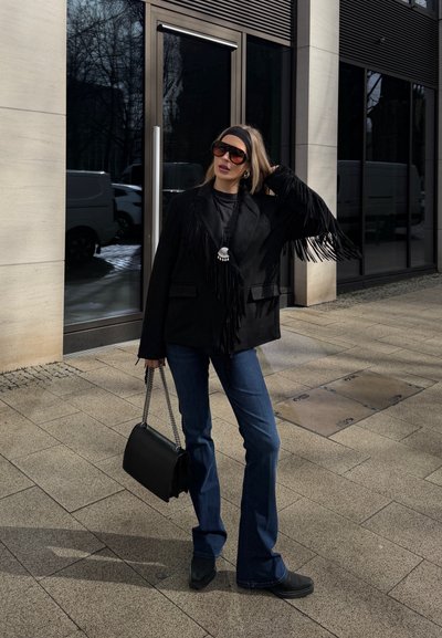 Mujer con chaqueta negra con flecos, gafas de sol oscuras, vaqueros azules acampanados, botas negras, diadema, sosteniendo un bolso negro en la acera de la ciudad.