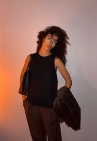 Personne aux cheveux bouclés portant un haut noir sans manches et un pantalon marron, tenant une veste marron et un sac noir, debout devant un mur clair.