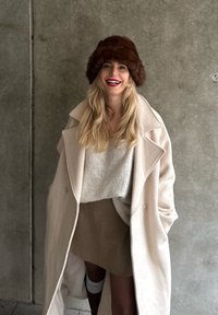 Long manteau en laine beige, pull en maille beige clair, jupe taupe, chapeau en fourrure marron, avec des matériaux texturés et une coupe décontractée. Posé devant un mur en béton.