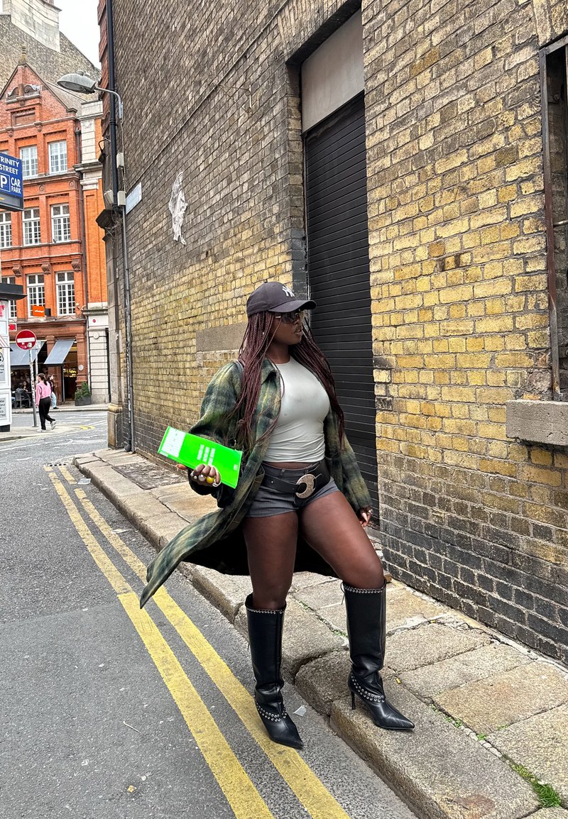 Plaid coat in green and black tones, grey top, black shorts, knee-high leather boots with silver accents, holding a bright green booklet.