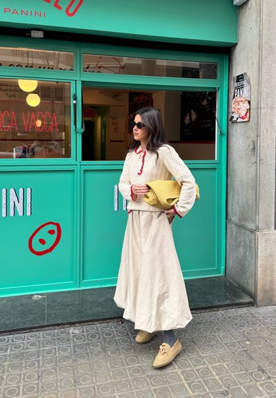 Una mujer está de pie frente a una tienda de color verde azulado, vistiendo un suéter de textura crema con acentos rojos y una larga falda beige. Ella sostiene una bolsa amarilla.