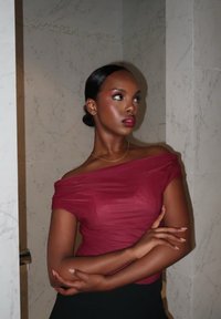 Femme avec chignon lisse portant un haut bordeaux à épaules dénudées, bras croisés, regardant sur le côté devant un fond de mur en marbre clair.