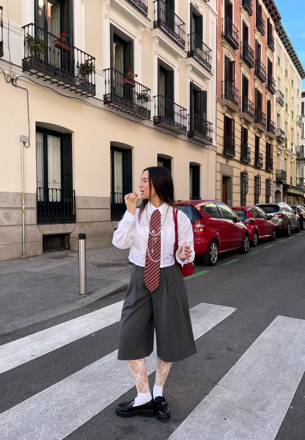 Camisa branca com uma gravata riscada a vermelho, culotes cinzentos e meias-calças com padrão de renda. Sapatos pretos, segurando uma bebida, numa rua da cidade.
