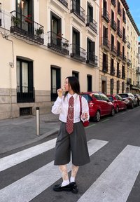 Camisa branca com uma gravata riscada a vermelho, culotes cinzentos e meias-calças com padrão de renda. Sapatos pretos, segurando uma bebida, numa rua da cidade.