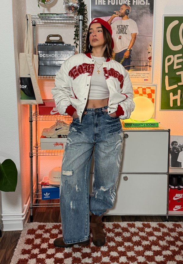 Mladá žena v červenej šiltovke, bielej krátkej bunde, voľných potrhaných džínsoch a hnedých topánkach stojaca v miestnosti s plagátmi a teniskami.