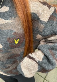 Camisola de malha camuflada cinzenta e bege com punho canelado e logótipo de pássaro amarelo no peito; textura macia com detalhes de padrão variados.