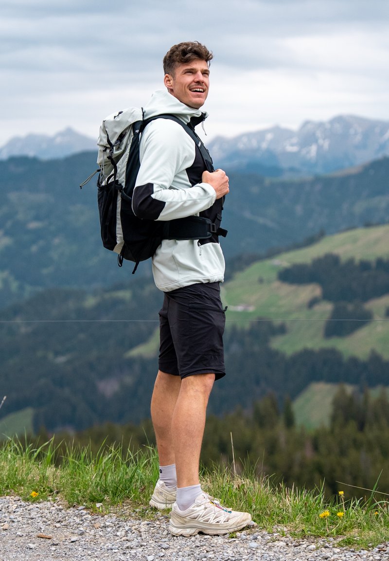 Homme portant un sac à dos et une tenue de sport, debout sur un sentier de montagne, souriant, avec des collines vertes au loin et un ciel nuageux en arrière-plan.