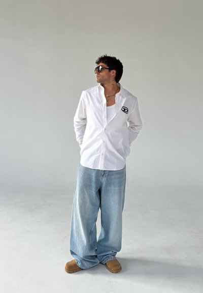 Camisa blanca de botones, corte relajado, mangas cortas, logo en el pecho; jeans oversize azul claro; zuecos marrones; gafas de sol; fondo neutro.