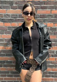 Jachetă din piele neagră cu fermoare argintii, top scurt negru și curea cu accente metalice. Fundal cu un zid din cărămidă roșie și gri.