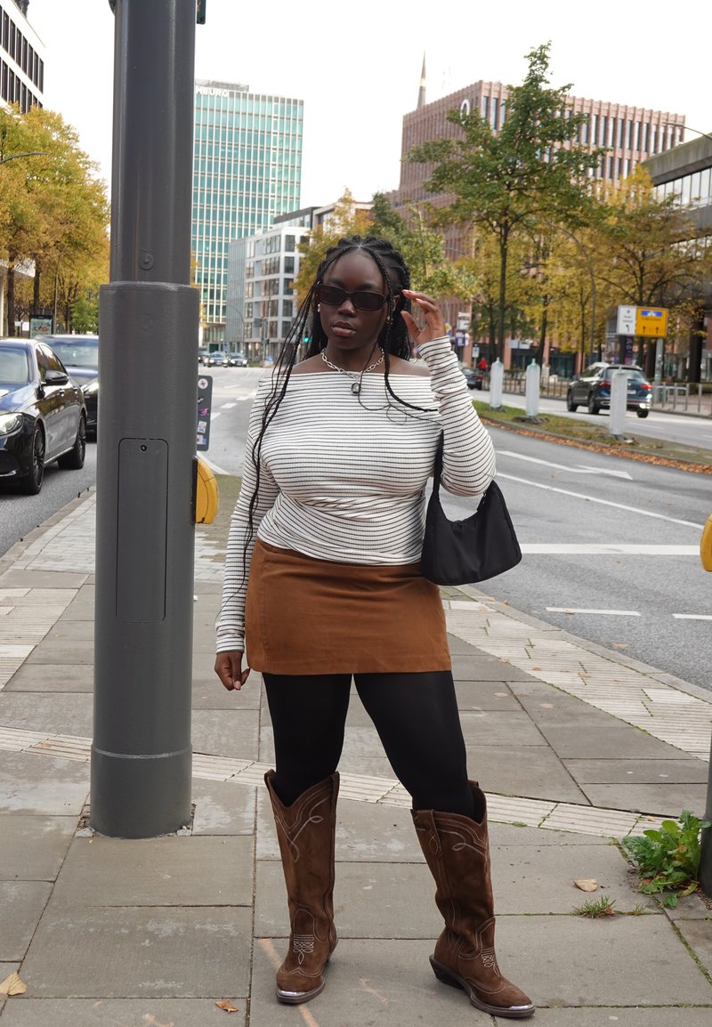 Haut à manches longues rayé, jupe en daim marron, leggings noirs, et bottes de cowboy marron avec des détails de couture. Sac à main noir et lunettes de soleil.