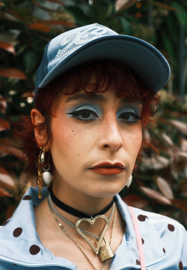 Osoba o krótkich czerwonych włosach nosi niebieską czapkę, wyrazisty niebieski cień do powiek, pomarańczową szminkę, kurtkę w kropki oraz warstwowe naszyjniki, w tym z kłódką w kształcie serca.