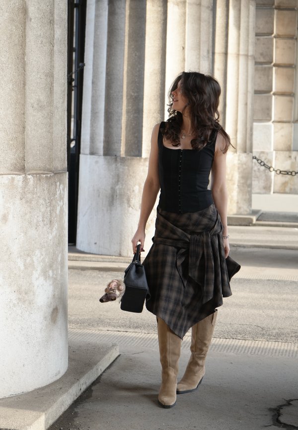 Nainen tummat kiharat hiukset, musta yläosa, ruudullinen hame ja beiget saappaat, pitää mustaa käsilaukkua, kävelee kivisärkkien vieressä.