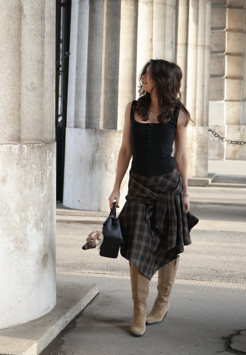 Kobieta o ciemnych kręconych włosach, ubrana w czarną bluzkę, kratowaną spódnicę i beżowe botki, trzymająca czarną torebkę, idąca obok kamiennych kolumn.