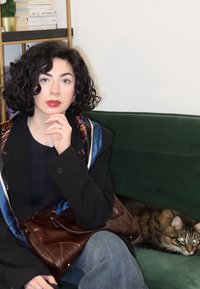 En person med krøllet sort hår iført en sort blazer, der holder en brun håndtaske, sidder på en mørkegrøn sofa ved siden af en tigerstribet kat.