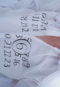 Vit skjorta med knappar och med djärva svarta siffror och symboler tryckta på ärmarna. Tyget har en subtil textur.