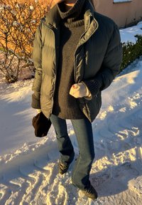Puffa jacket in un verde scuro con zip e chiusura a scatto, maglione a collo alto grigio, jeans a zampa d'elefante blu e calzature nere, in un ambiente innevato.