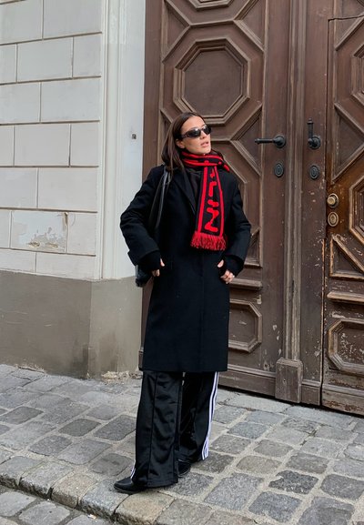 Abrigo negro y pantalones deportivos negros con rayas blancas. Bufanda roja con texto contrastante, gafas de sol, de pie frente a una puerta de madera.
