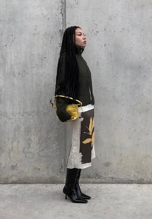 Mujer con trenzas largas de pie contra una pared gris, lleva un suéter verde oscuro, falda con estampado, botas negras de tacón y sostiene un pequeño bolso amarillo.
