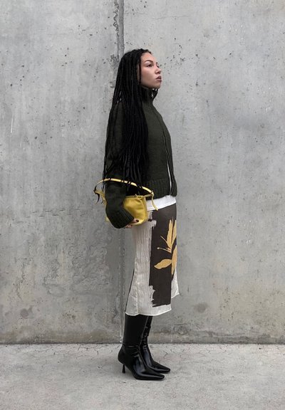 Mujer con trenzas largas de pie contra una pared gris, lleva un suéter verde oscuro, falda con estampado, botas negras de tacón y sostiene un pequeño bolso amarillo.