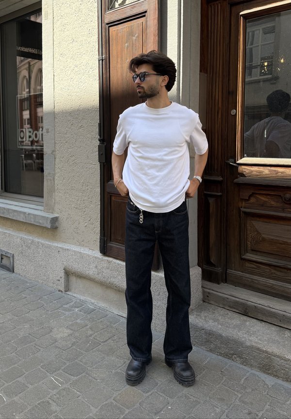 Mann trägt weißes T-Shirt, dunkle Jeans, schwarze Stiefel und Sonnenbrille und steht auf gepflasterter Straße nahe hölzerner Türen.